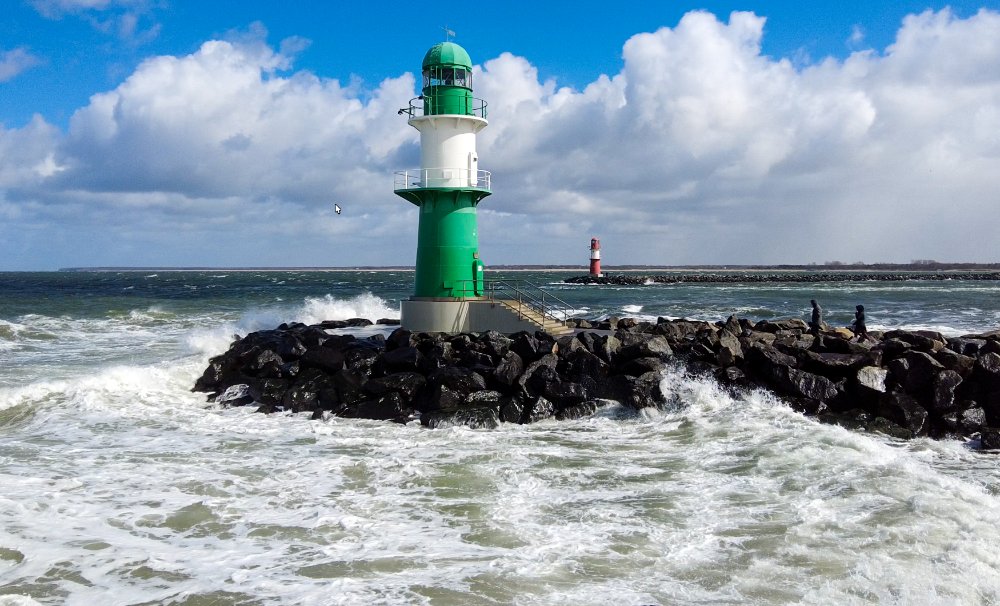 The green-and-white pier light of the Warnemünde west pier in a stormy Baltic Sea, waves crashing against the pier head under a partly cloudy blue sky, with the red east pier light in the background.