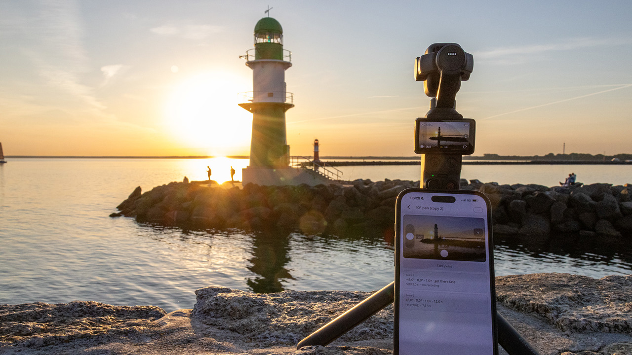An Osmo Pocket on a tripod films the pier light of Warnemünde's west pier at sunrise; in front of it an iPhone running Pathset with a camera move for a 90-degree pan.
